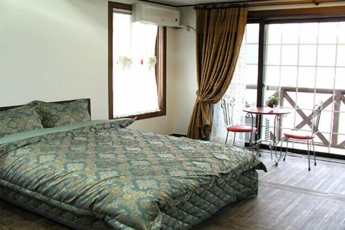 Een slaapkamer met een groot bed, een raam met gordijnen, en een balkon met stoelen en een tafel.