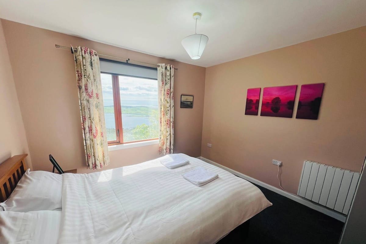 A hotel room with a bed, a window with curtains, a radiator, and two paintings on the wall.