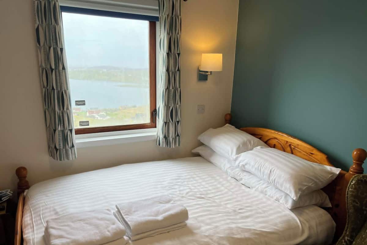 A neatly made bed with white linens and a folded towel, a wooden headboard, a window with curtains, and a wall-mounted light fixture.