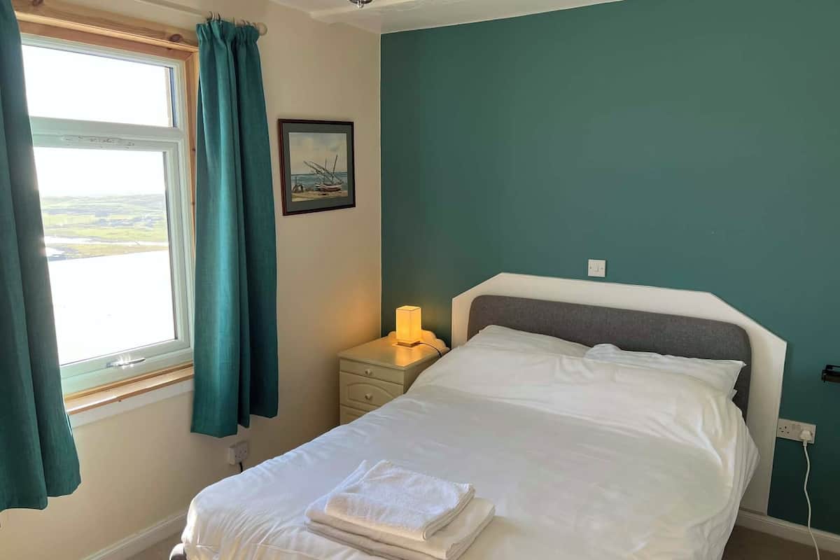 A bedroom with a bed, a nightstand, a lamp, a window with curtains, and a painting on the wall.