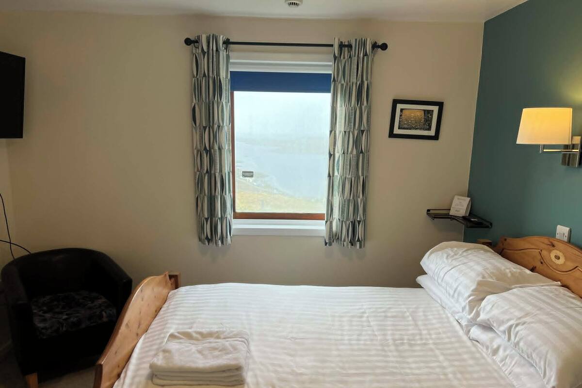 A hotel room with a bed, a chair, a window with curtains, a picture on the wall, and a lamp.