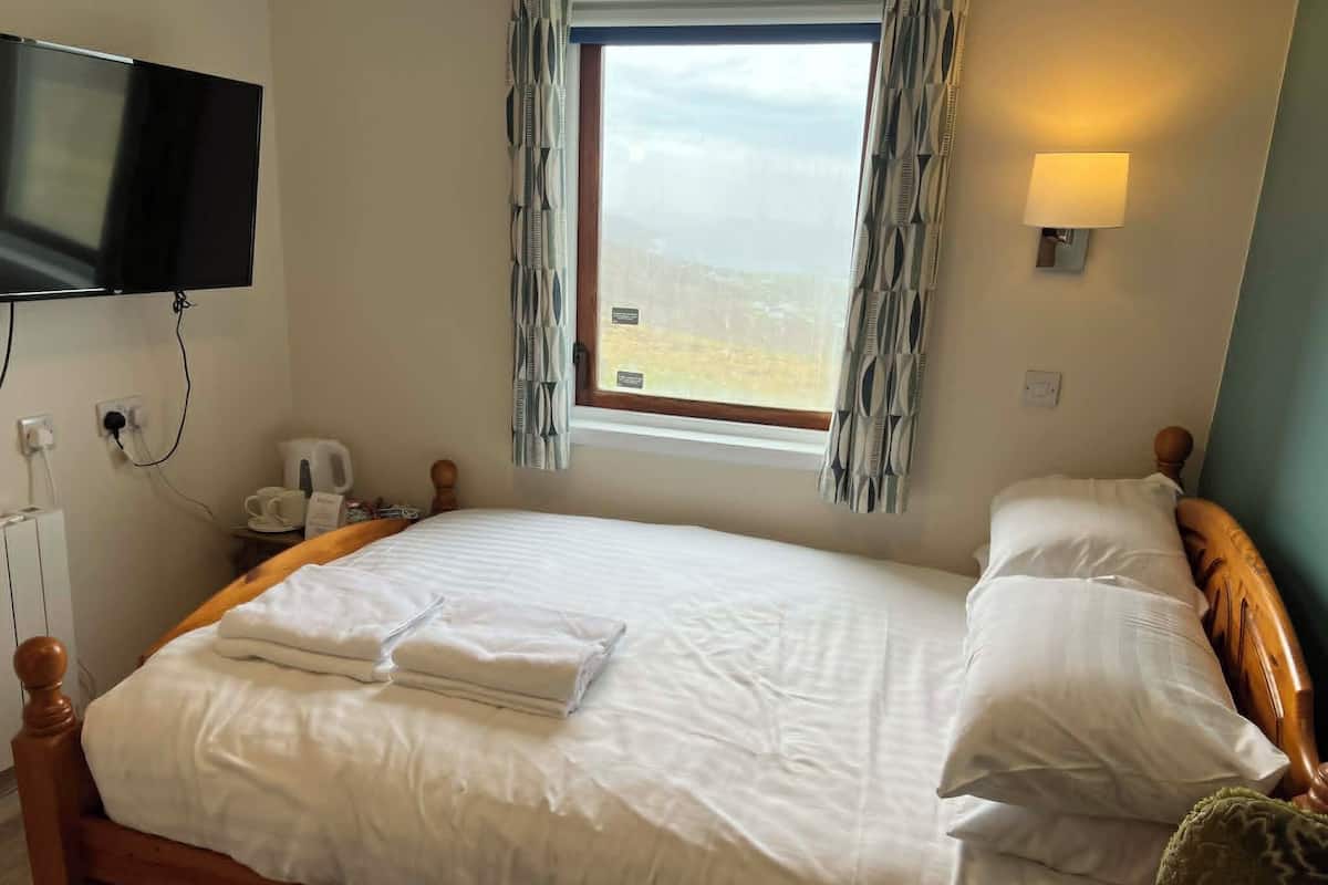 A neatly made bed with white linens, a bedside table with a lamp, a television mounted on the wall, and a window with curtains.