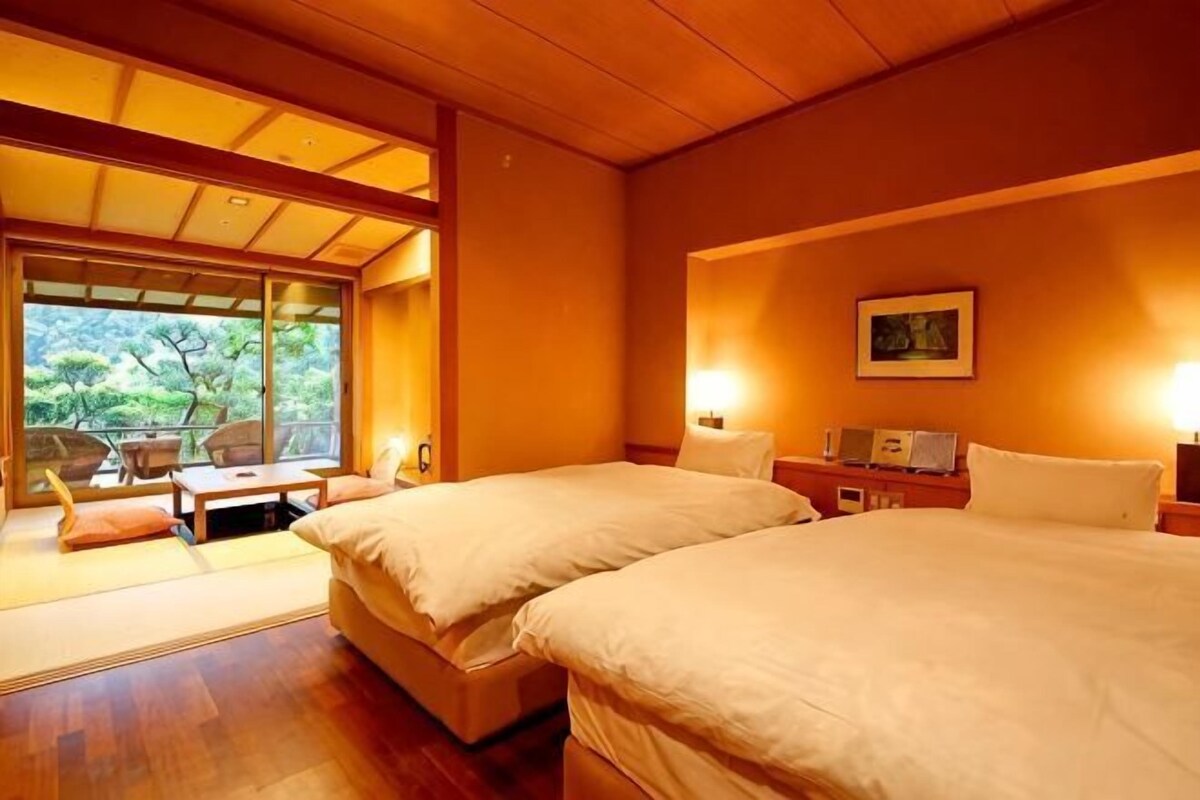 A hotel room with two beds, a sitting area with a table, and a large window offering a view of greenery.