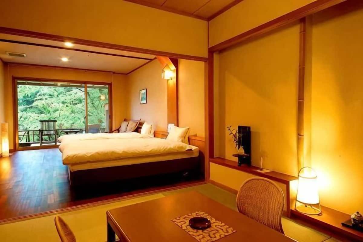 Una camera da letto con un letto, un tavolo e una sedia. Si vede della vegetazione attraverso la finestra.