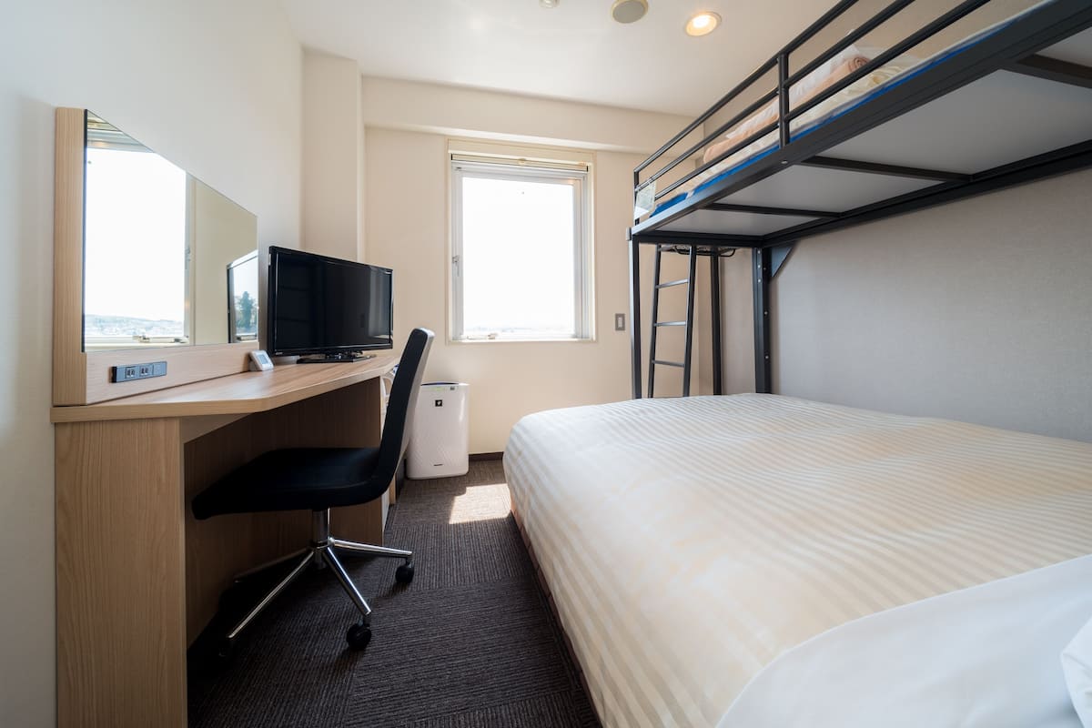Ein Hotelzimmer mit einem Etagenbett, einem Schreibtisch mit Computer, einem Stuhl und einem Fenster.