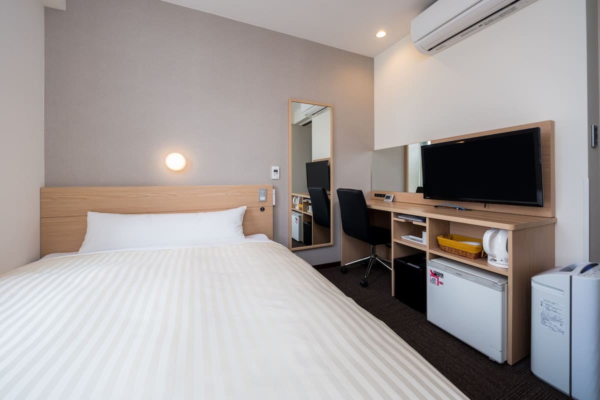 Ein Hotelzimmer mit Bett, Schreibtisch, Stuhl, Fernseher, Spiegel, kleinem Kühlschrank und Klimaanlage.