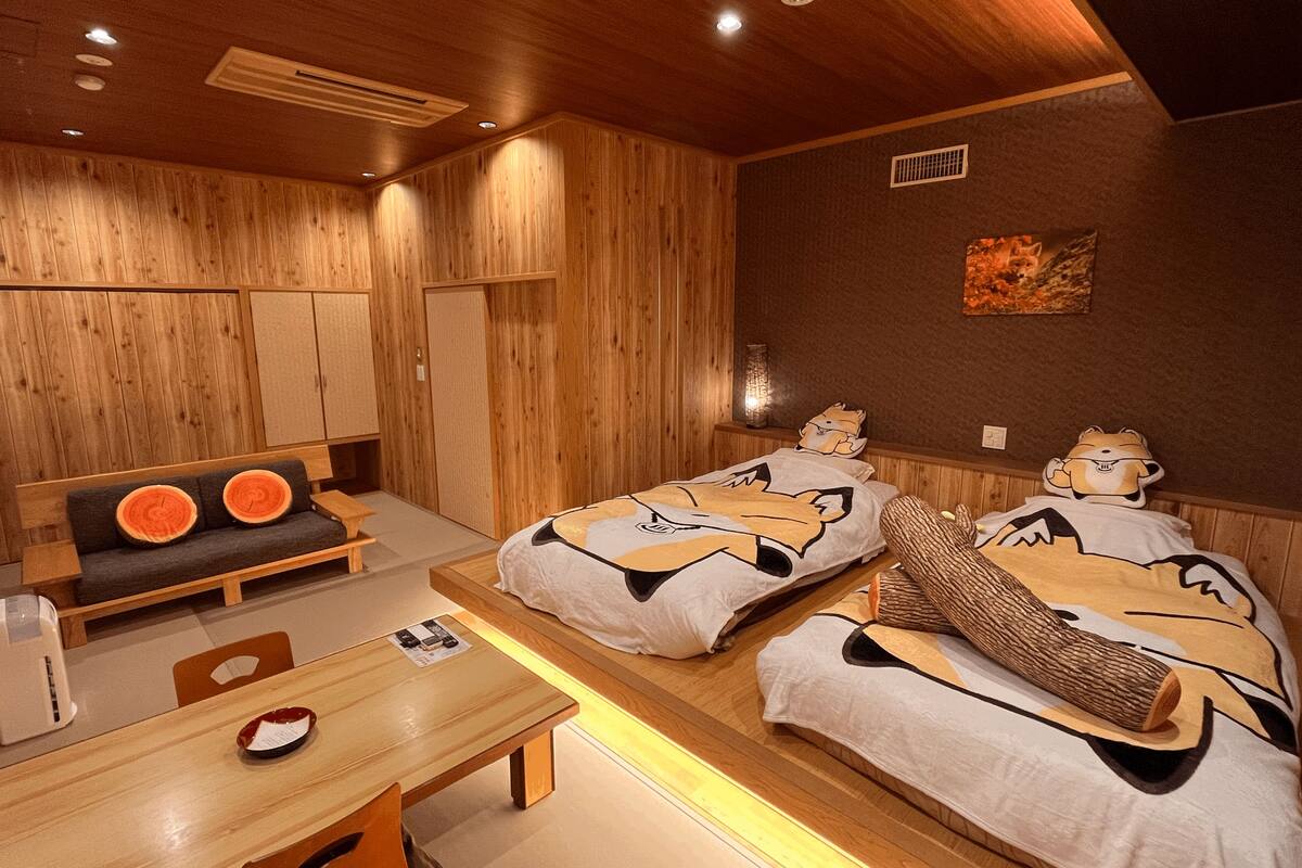 A room with wooden walls and ceiling, two beds with cartoon character bedding, a wooden bench, a table, and a small shelf with a decorative item.