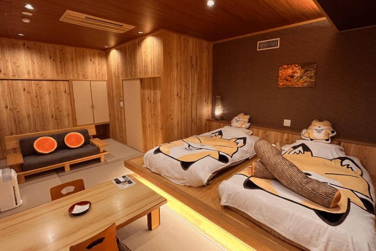 A room with wooden walls, a sofa with orange cushions, two beds with cartoon character bedding, a wooden table, and a small table with a red and white object on it.