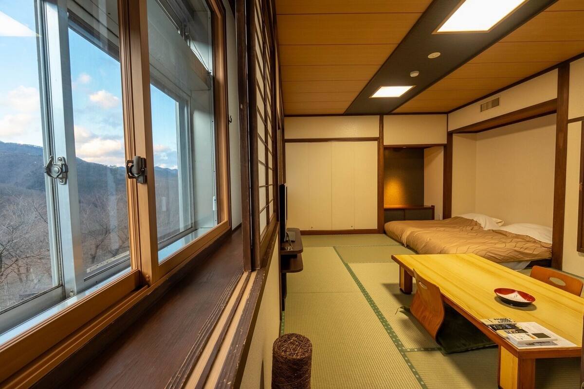 A traditional Japanese room with tatami flooring, a futon bed, a low wooden table, and a view of mountains through large windows.