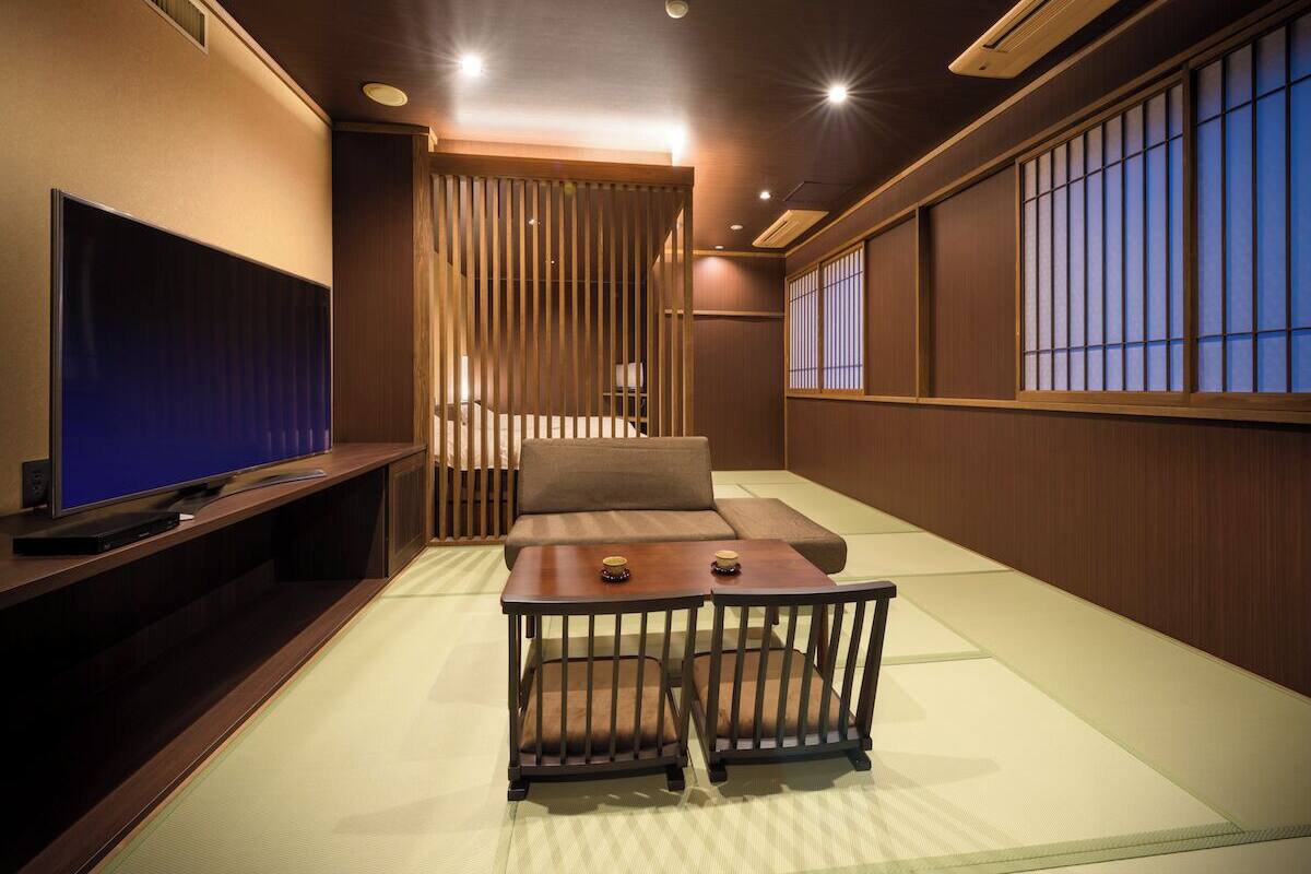 A traditional Japanese room with tatami flooring, a low table with cushions, a television on a stand, and sliding doors.