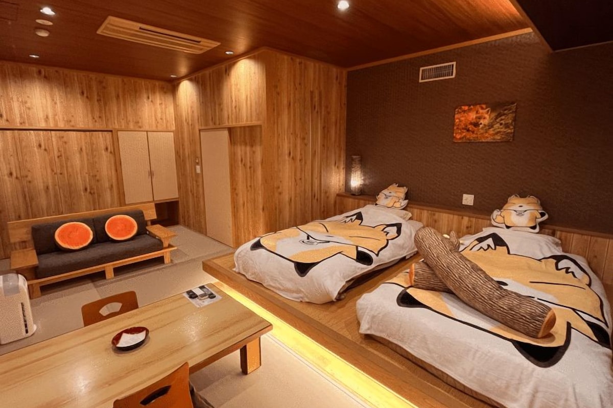 A room with wooden walls, a sofa with orange cushions, two beds with cartoon character bedding, a wooden table, and a small table with a red and white object on it.