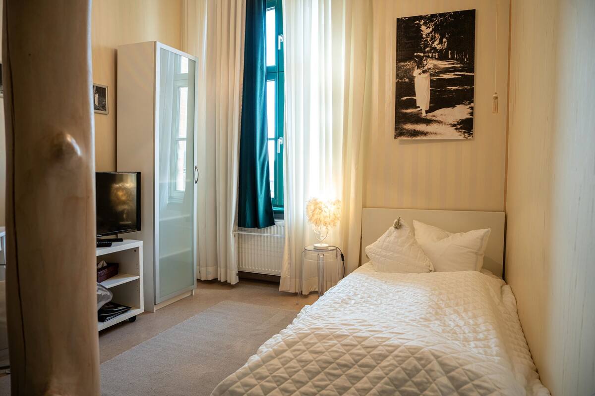 A bedroom with a bed, a television on a stand, a wardrobe, and a window with curtains.
