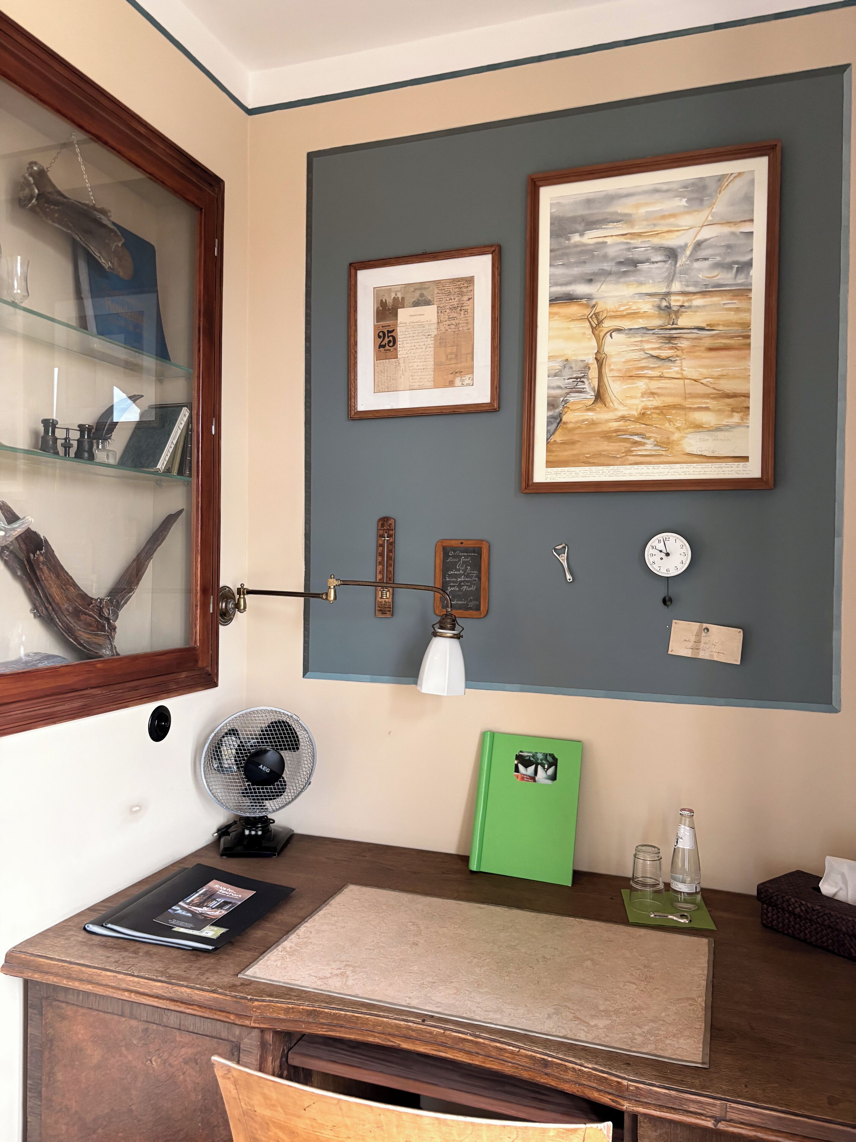 Una habitación con un escritorio de madera, una carpeta verde, un reloj y obras de arte enmarcadas en la pared.