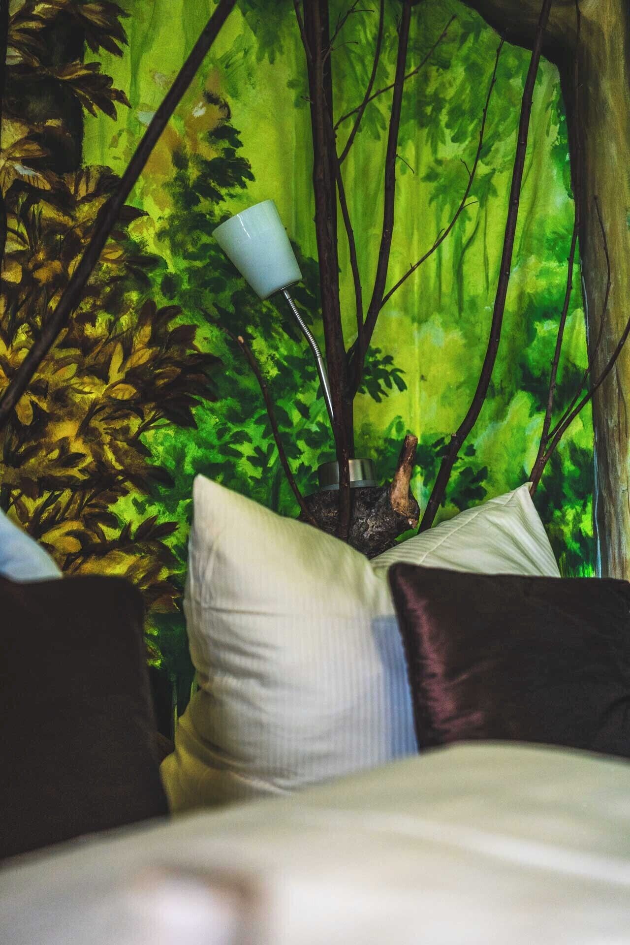 Ein Bett mit Kissen und einer Lampe in einem Raum, dessen Wand ein Waldmotiv zeigt.