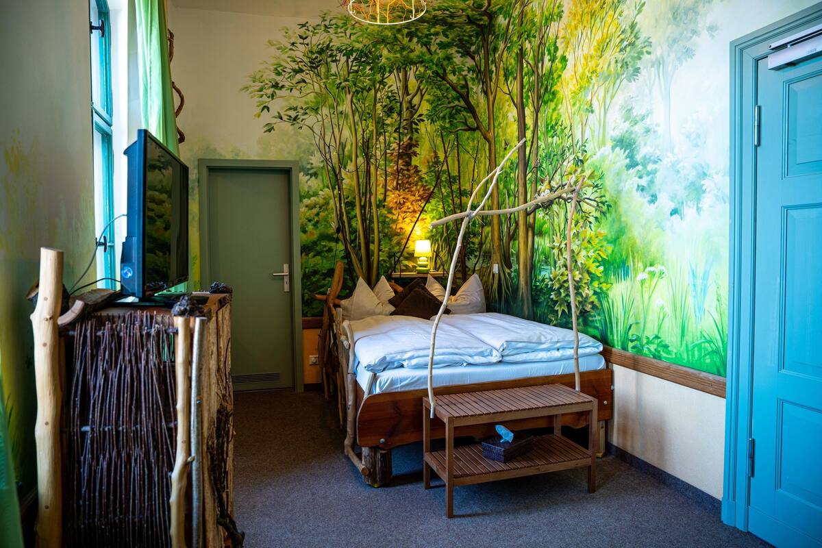 A bedroom with a bed, a nightstand, a lamp, a wall mural, and a door.