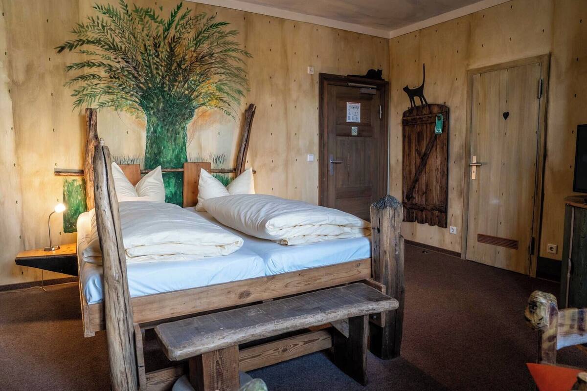 A rustic wooden room with a bed, a bench, a large plant, and a cat sculpture.