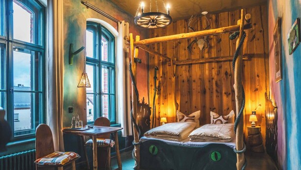 A rustic room with a four-poster bed, wooden walls, a dining table with chairs, and a view through a large window.