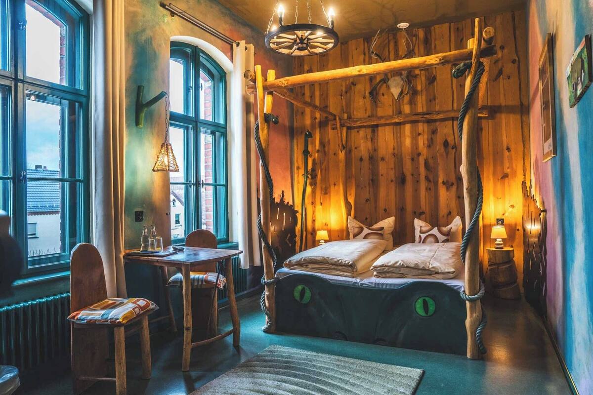 A rustic room with a four-poster bed, wooden walls, a dining table with chairs, and a view through a large window.