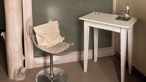 A room with a glass chair, a small white table with a bottle and glass, and a large window with a view of buildings.