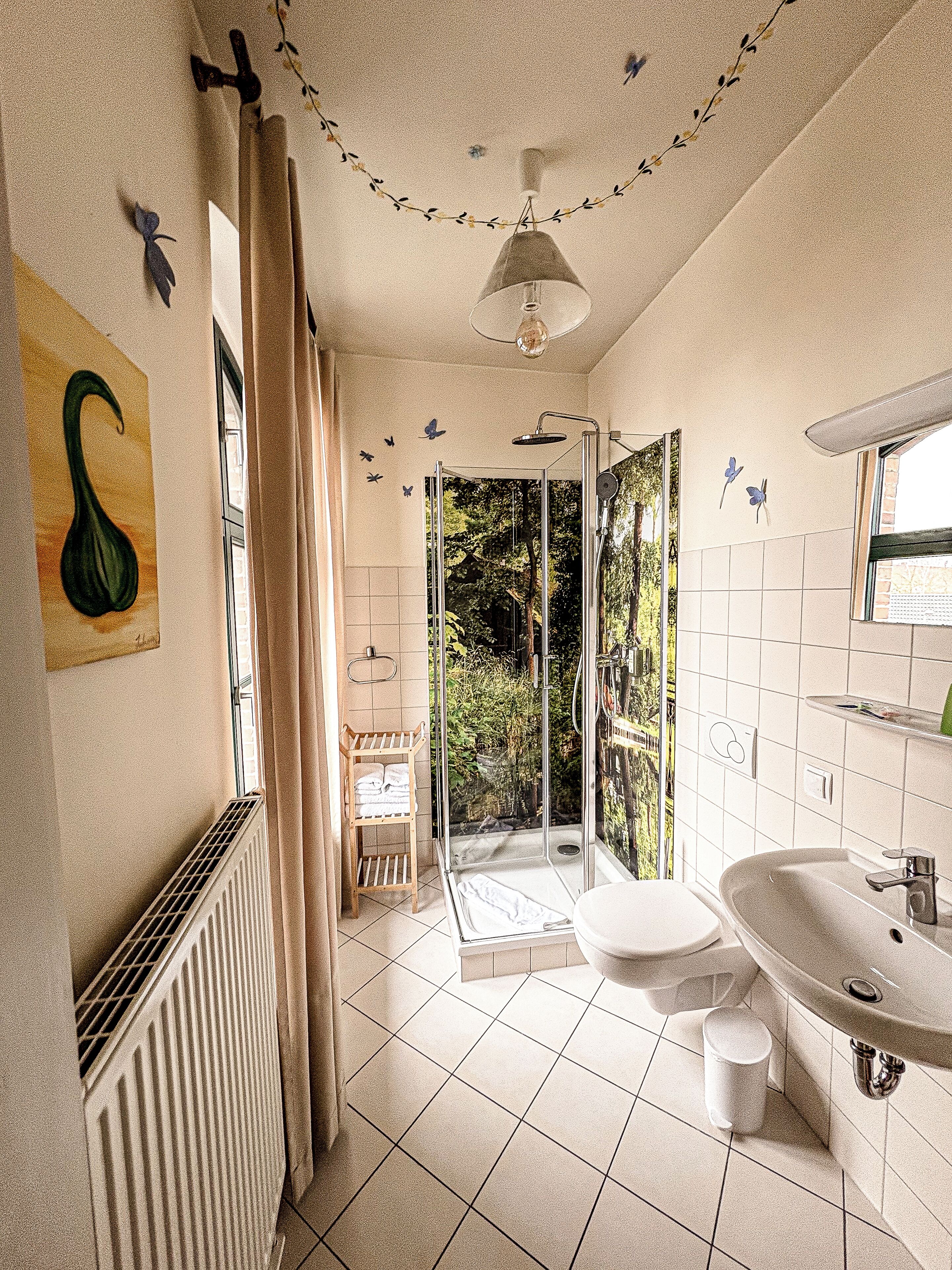 Un baño con ducha, inodoro y lavamanos. Hay un radiador, un perchero para toallas y una ventana con cortinas.