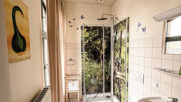 Un baño con ducha, inodoro y lavamanos. Hay un radiador, un perchero para toallas y una ventana con cortinas.