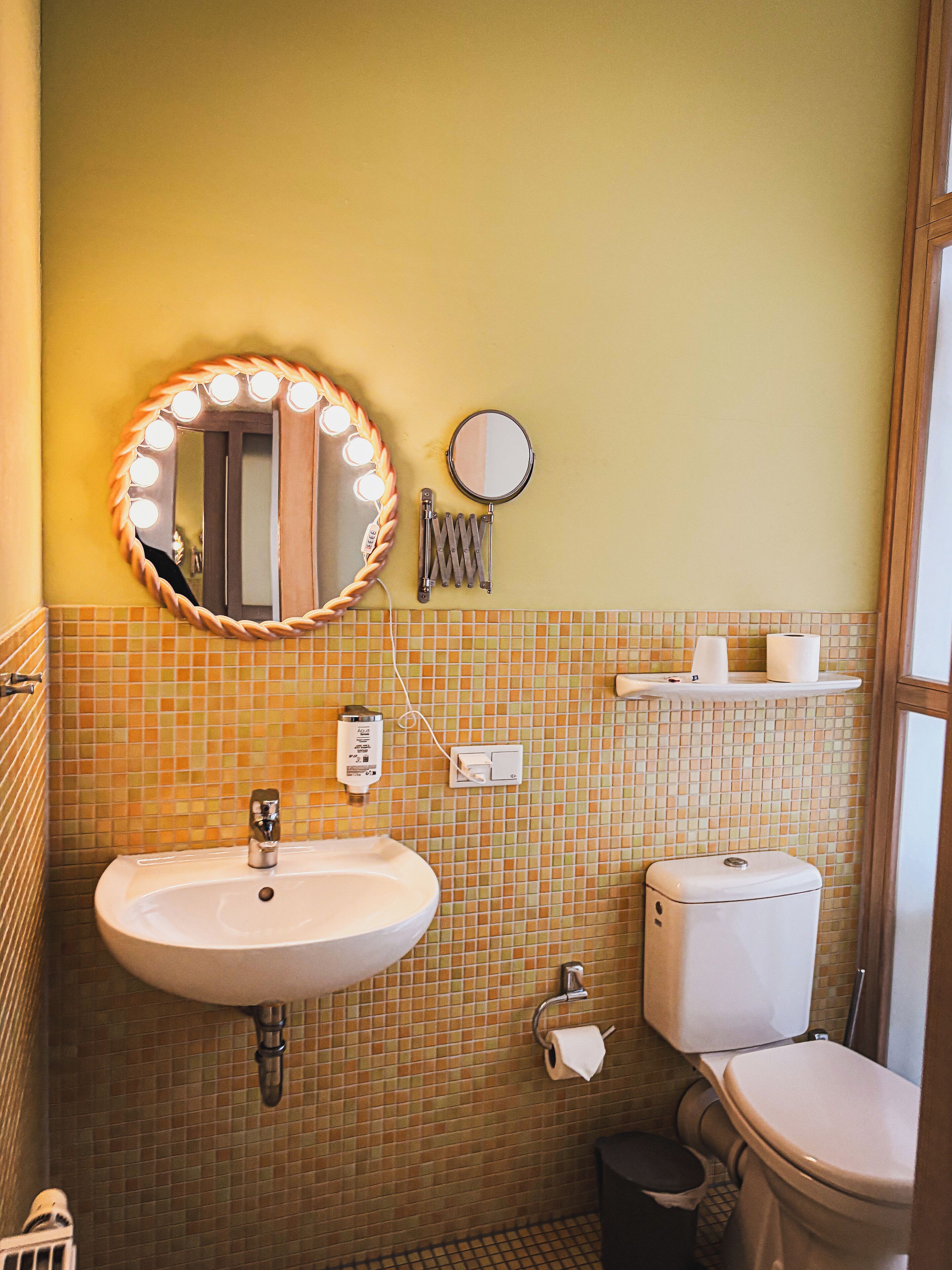 Une salle de bain équipée d’un lavabo, d’un miroir, de toilettes et d’une petite étagère où sont rangés les articles de toilette.