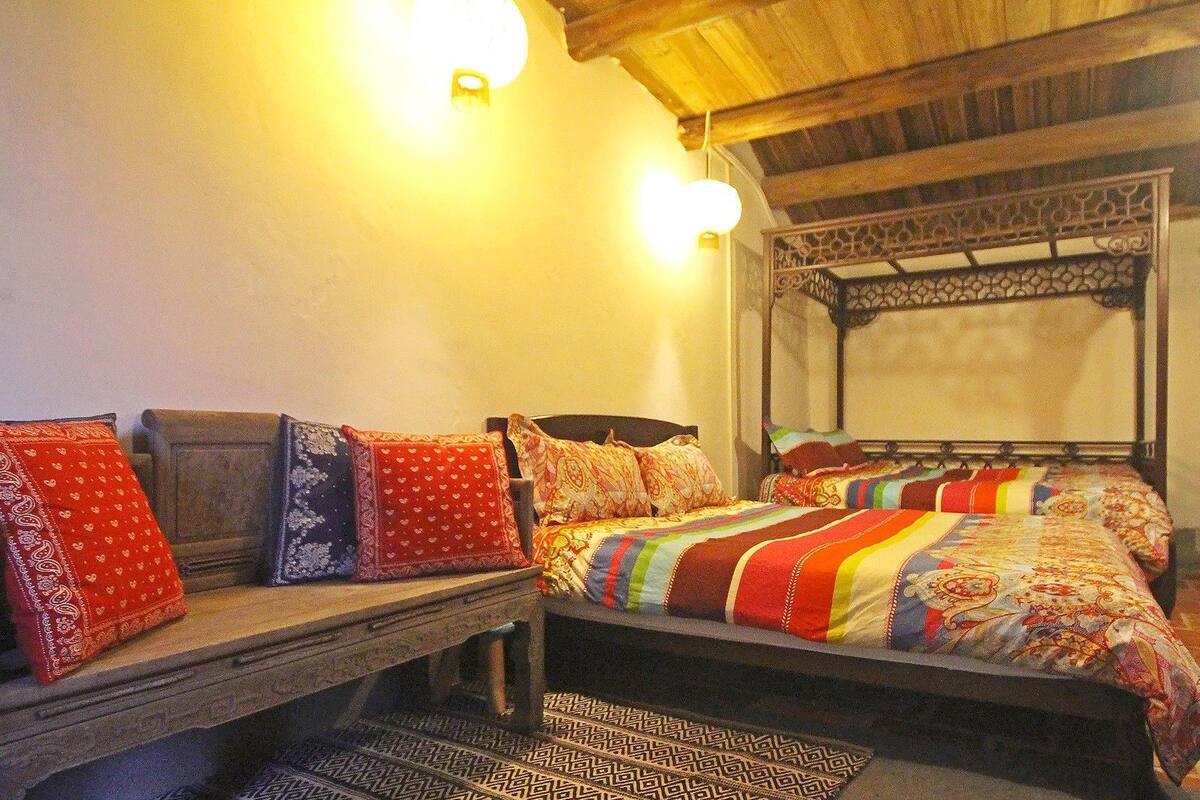 Una cama con dosel tradicional, con un colcha de colores, un banco de madera con cojines y una alfombra estampada.