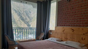 A hotel room with a large bed, a wooden headboard, a view of a mountainous landscape, and a balcony with a chair. - White Water Inn McLeodganj (Dharamshala)