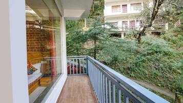 A balcony with a wooden floor and a white railing, overlooking a lush green area with trees and a building in the background.