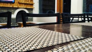 A dining area with a wooden table and chairs, a woven placemat, and a TV mounted on a stone wall. - White Water Inn McLeodganj (Dharamshala)