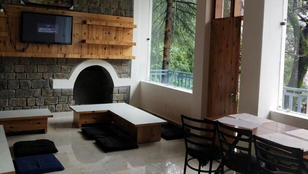 A cozy room with a stone fireplace, wooden shelves, a TV, and a large window offering a view of trees. - White Water Inn McLeodganj (Dharamshala)