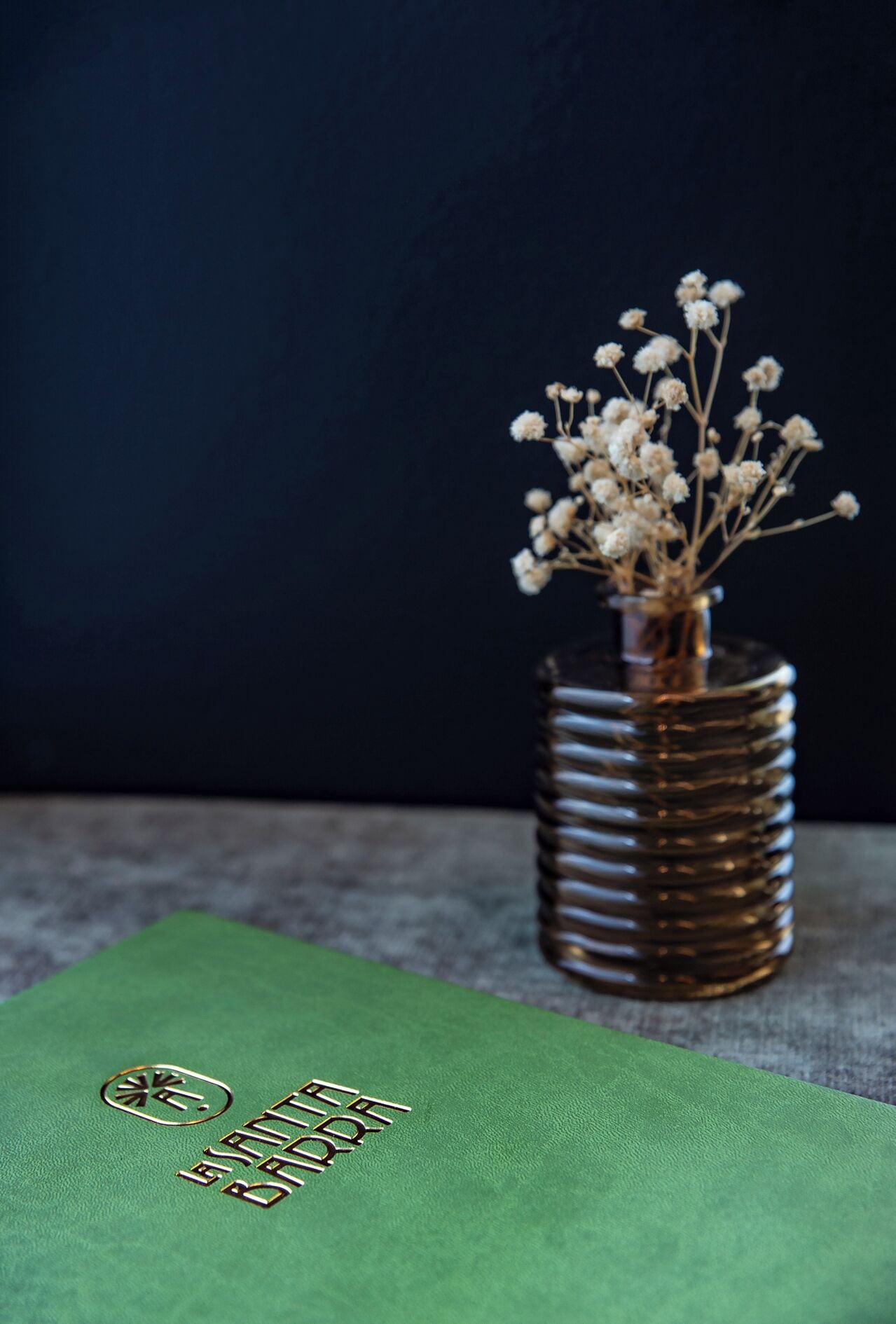 Ein grünes Buch mit geprägter Schrift, eine metallische Vase mit getrockneten Blumen und ein dunkler Hintergrund.