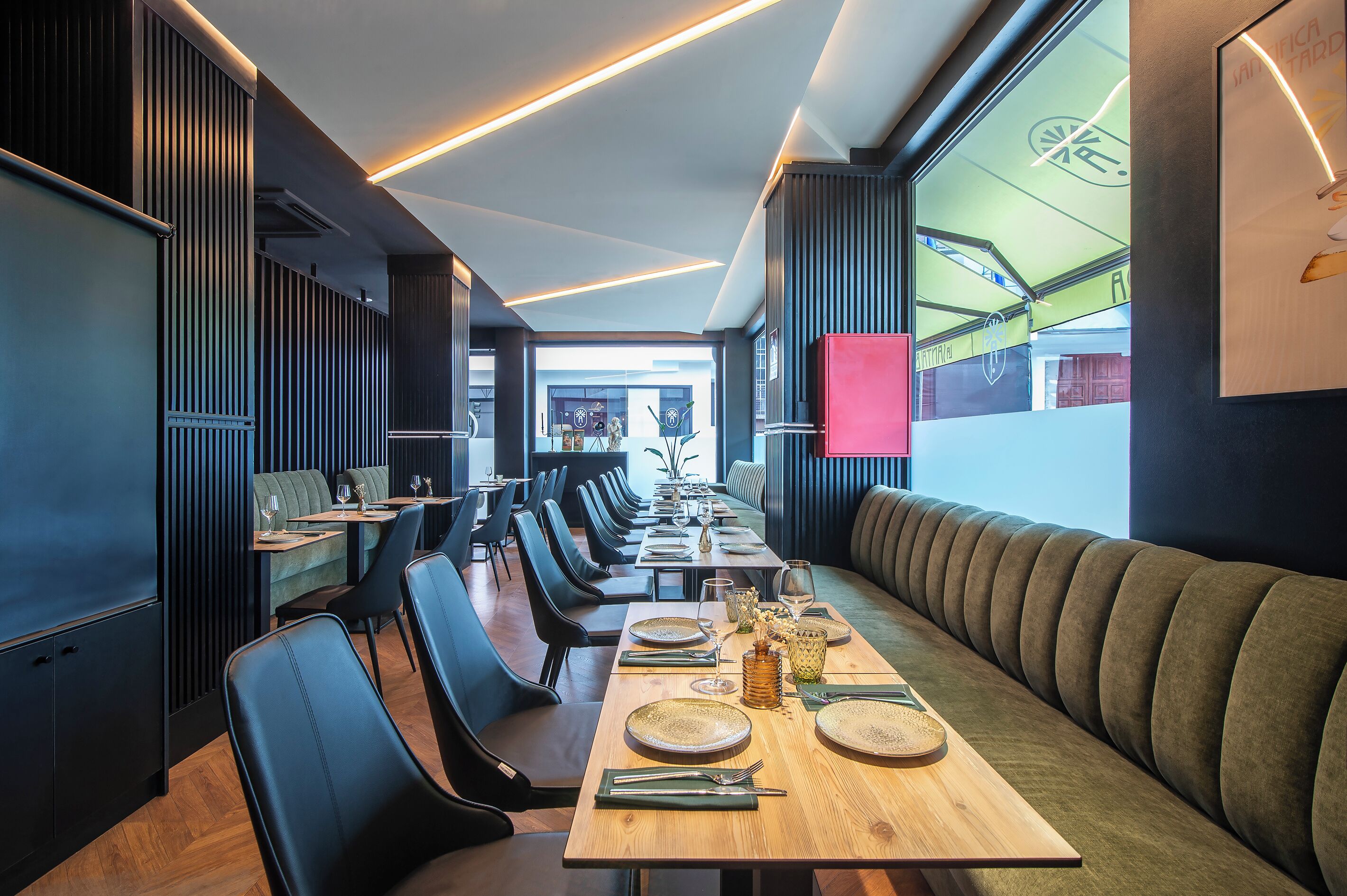 A modern restaurant with long communal tables, black upholstered chairs, and a wooden floor.