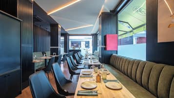A modern restaurant with long communal tables, black upholstered chairs, and a wooden floor.