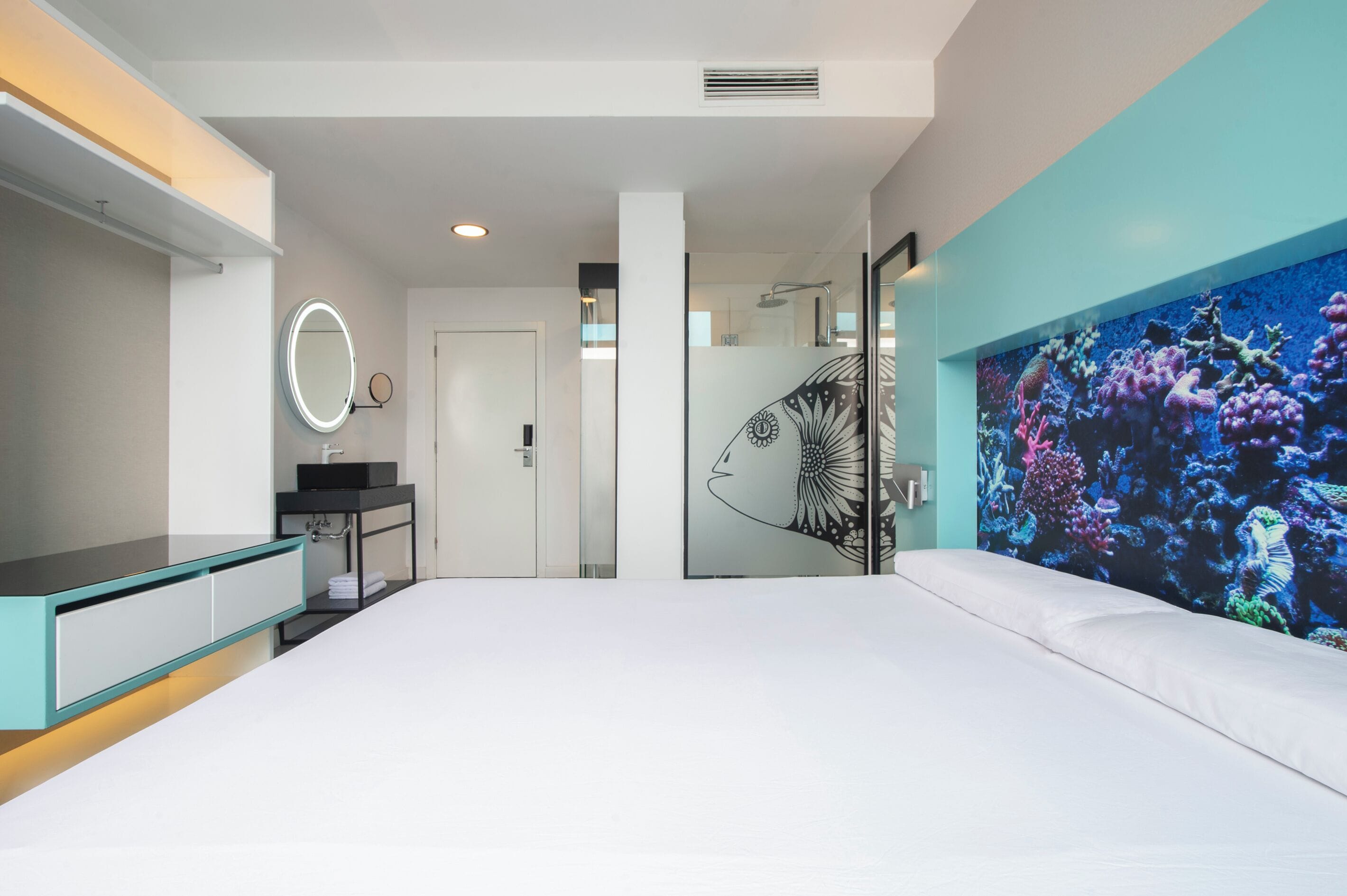 Una habitación de hotel moderna con una cama grande, un baño con espejo y lavamanos, y un mural vibrante con un arrecife de coral.