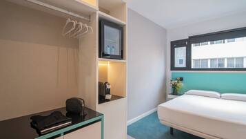 A hotel room with a bed, a desk with a chair, a shelf with toiletries, and a window with a view of buildings.