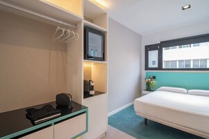 A hotel room with a bed, a desk with a chair, a shelf with toiletries, and a window with a view of buildings.