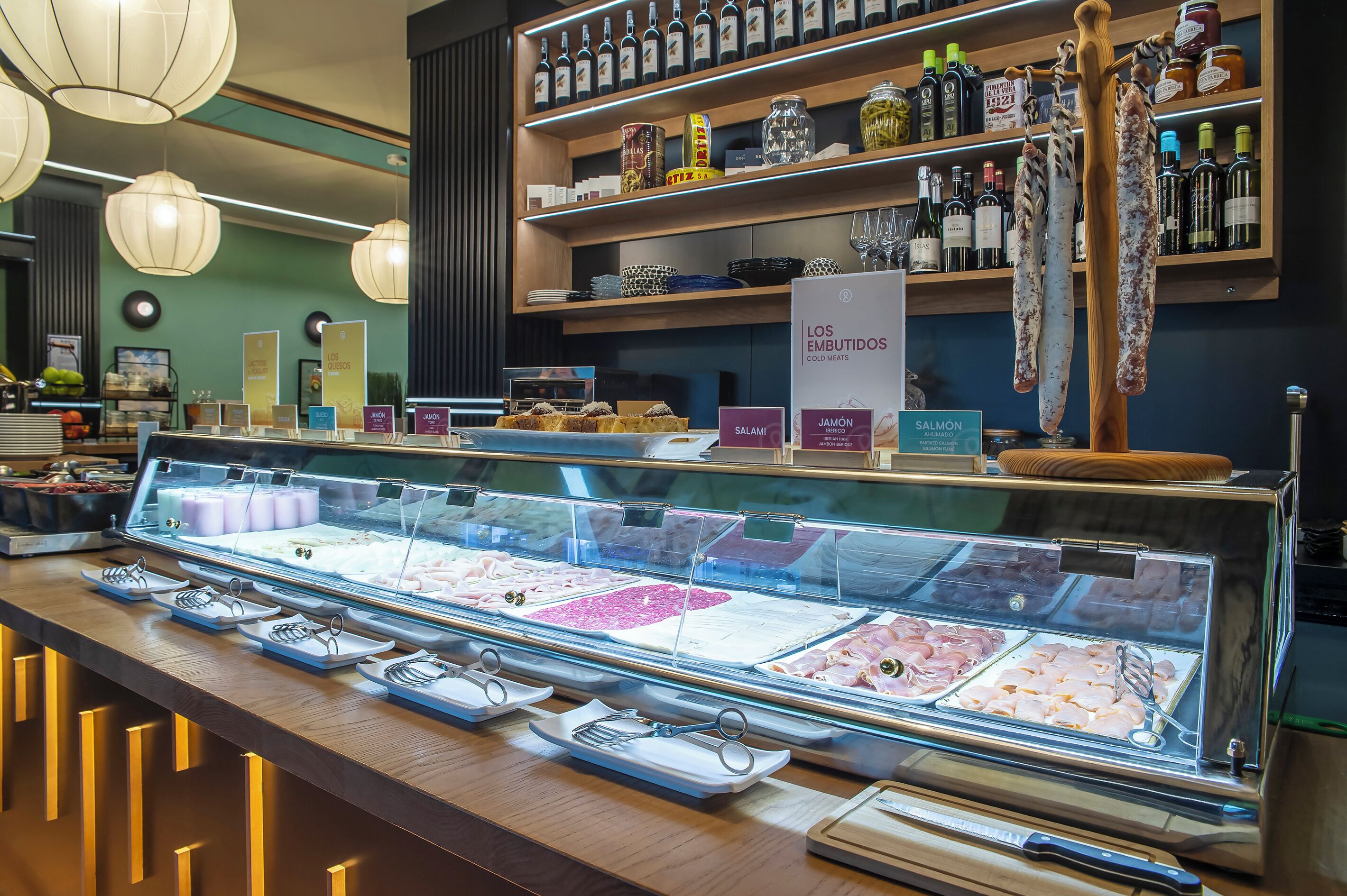 Mostrador de delicatessen con diversos tipos de embutidos y quesos, una estantería con vino arriba y un letrero que indica fiambres.