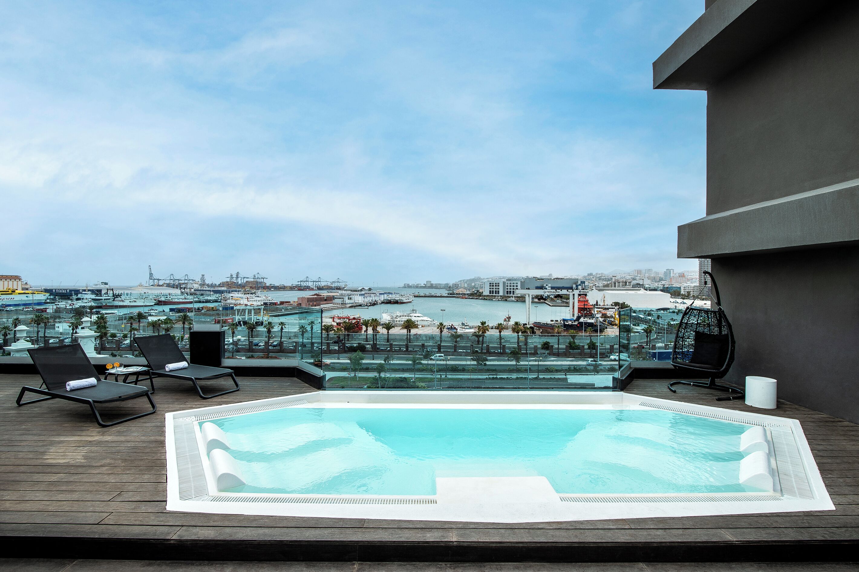 A rooftop pool with lounge chairs and a glass balustrade overlooking a marina and cityscape.