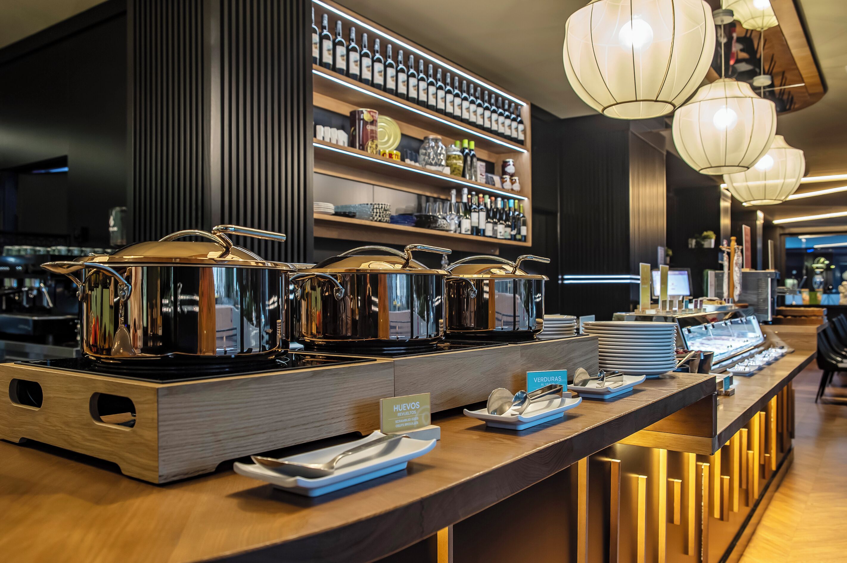Un espace buffet doté de plusieurs points de service, dont un espace intitulé « HUEVOS » pour les œufs, et une variété de bouteilles et de bocaux disposés sur des étagères à l’arrière-plan.