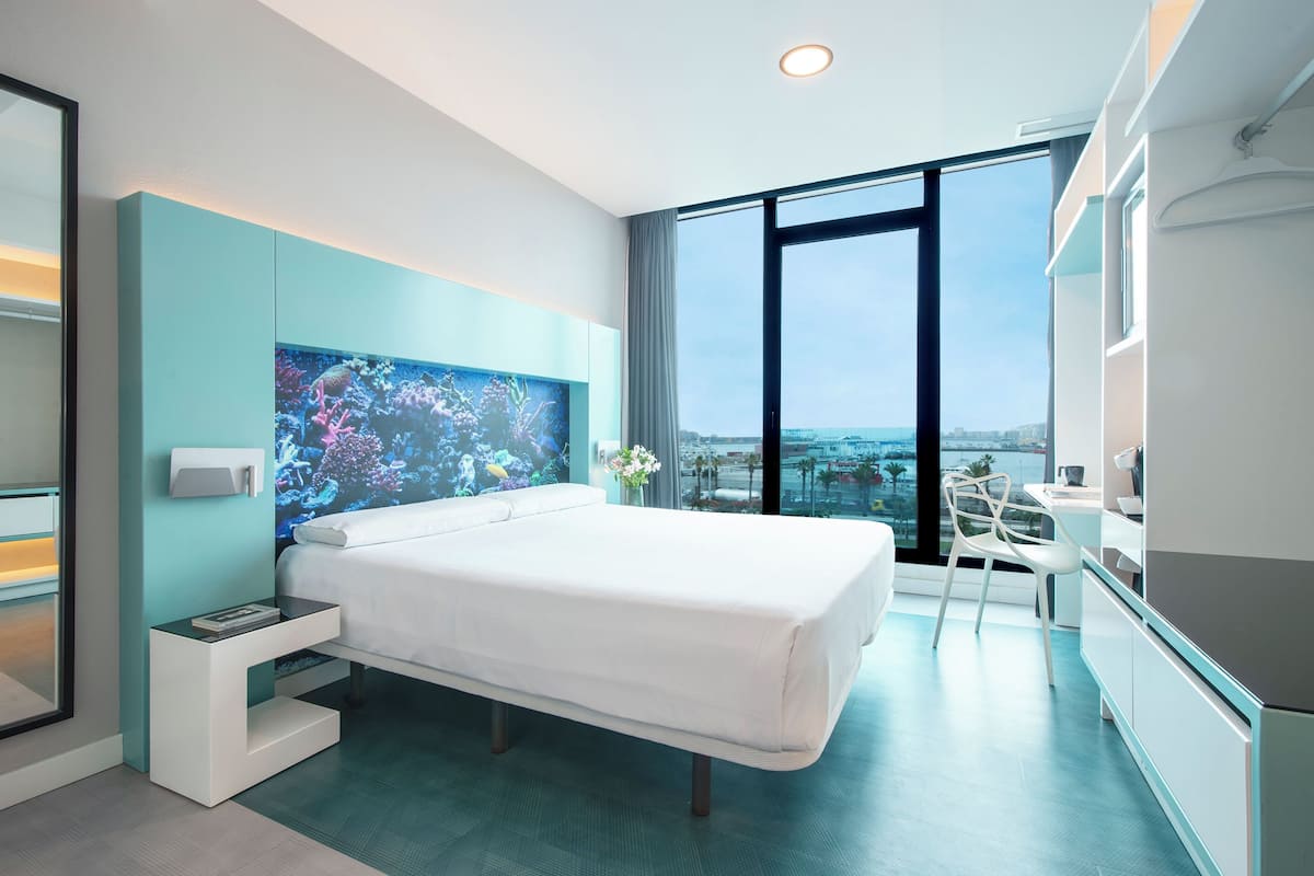 A modern hotel room with a large bed, a TV, a desk, and a view of the city.