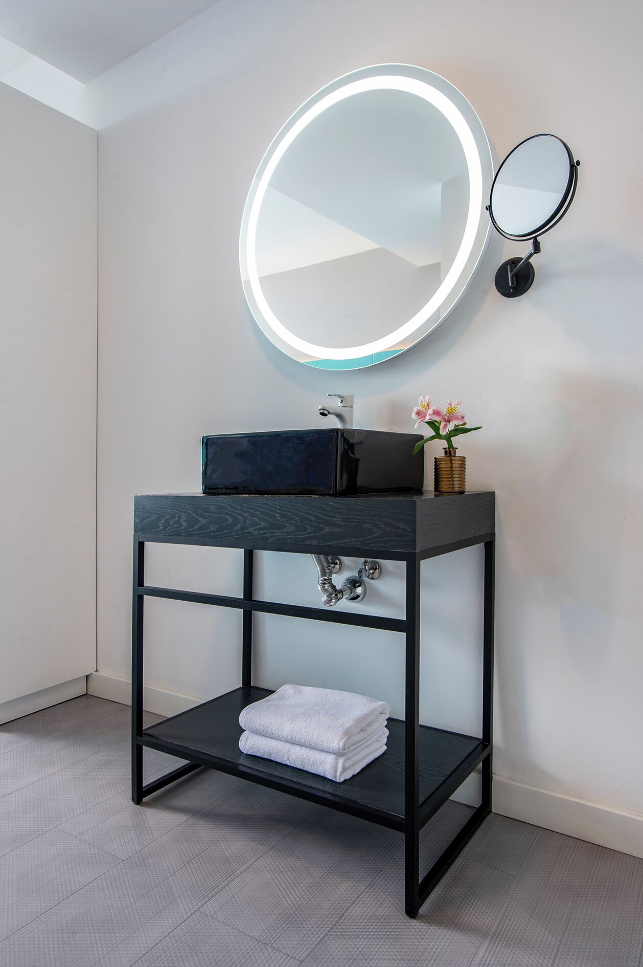 A bathroom with a black vanity, a round mirror with a light, and a small shelf with a towel.