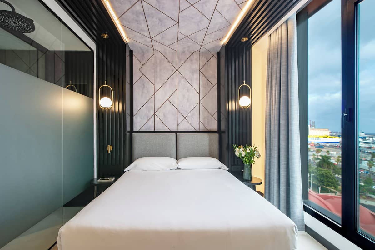 A modern hotel room with a large bed, two bedside tables, a nightstand, a wall-mounted light fixture, and a view of the city through a large window.