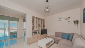 Un salón moderno con un sofá color beige, una mesa de centro blanca y un televisor sobre un mueble de madera. Cuenta con un balcón con vistas al mar.