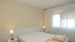 A hotel room with a large bed, two bedside tables, a lamp on each table, and a window with sheer curtains. - Apartment for 3 Persons in Quiet Part of Premantura With Beautiful Garden and Partial sea View (Medulin)