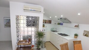 A compact kitchen with white cabinets, a gas stove, and a small sink. There is a wooden dining table with two chairs, a potted plant, and a decorative wall panel. - Apartment for 3 Persons in Quiet Part of Premantura With Beautiful Garden and Partial sea View (Medulin)