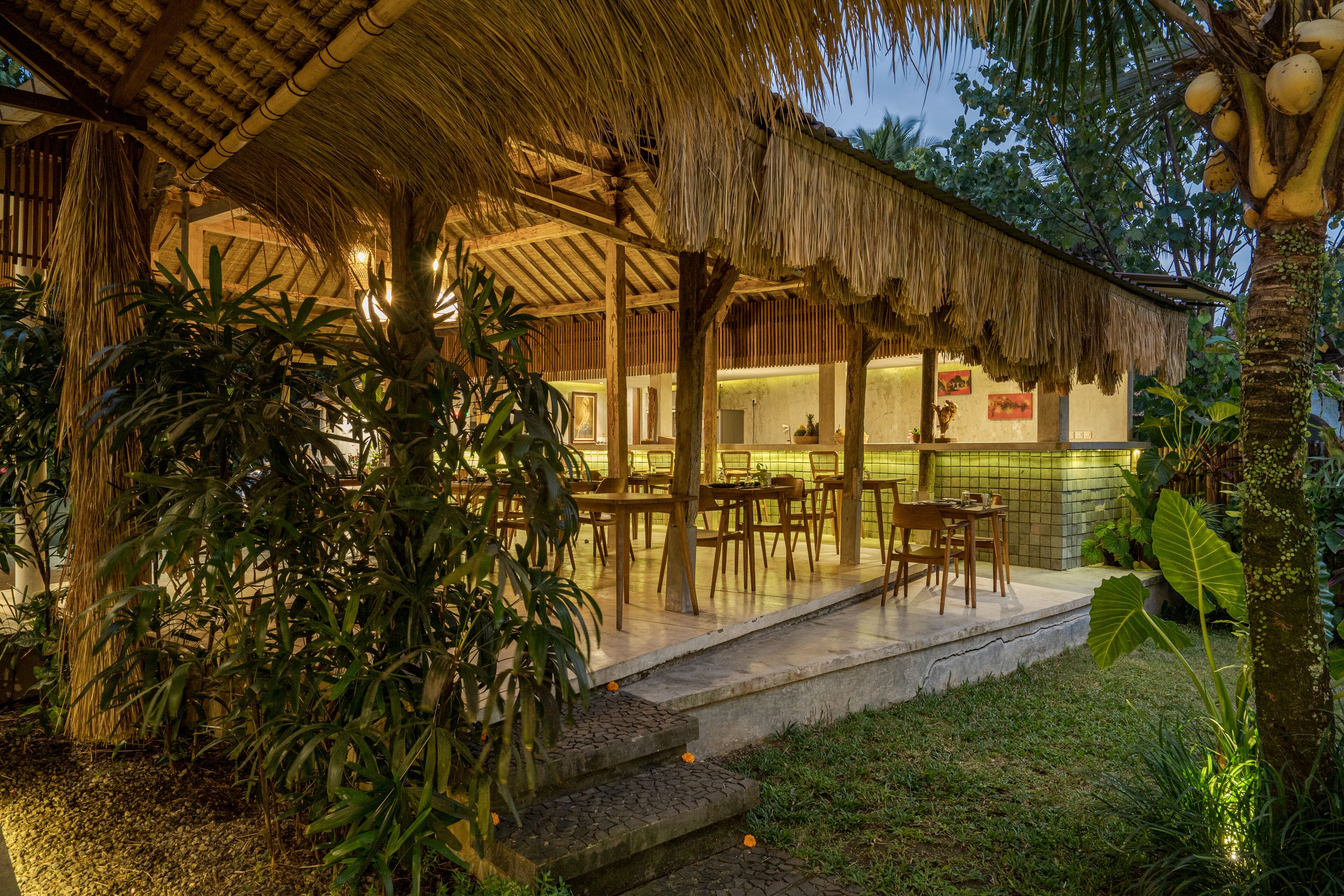 A thatched-roof outdoor dining area with tables and chairs, surrounded by lush greenery and a well-maintained garden.