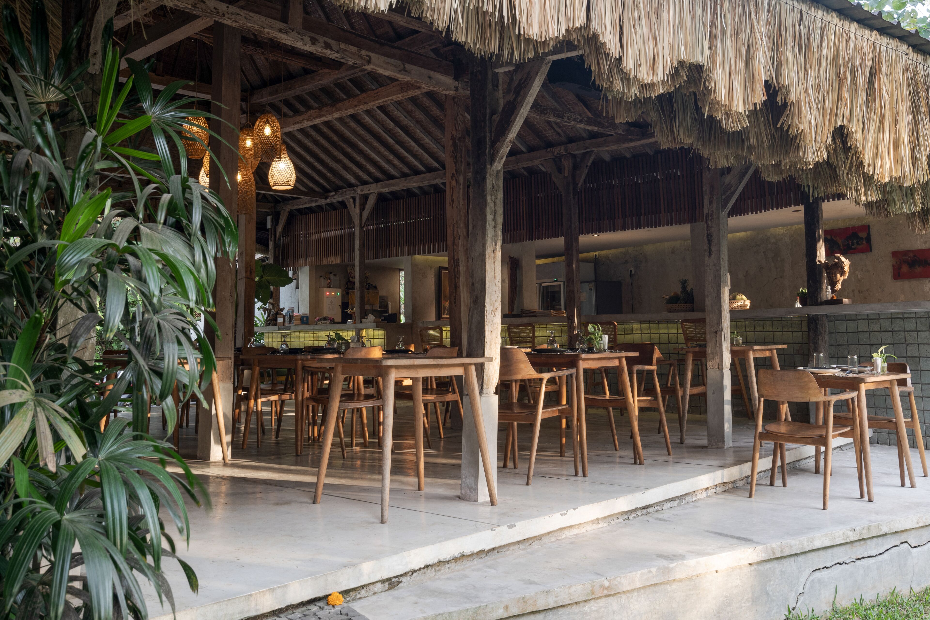 An open-air restaurant with wooden tables and chairs, a thatched roof, and indoor lighting.