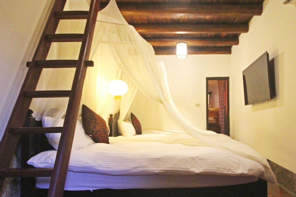 A bedroom with a wooden ladder leading to a bed with a canopy. There is a bedside lamp and a framed picture on the wall.