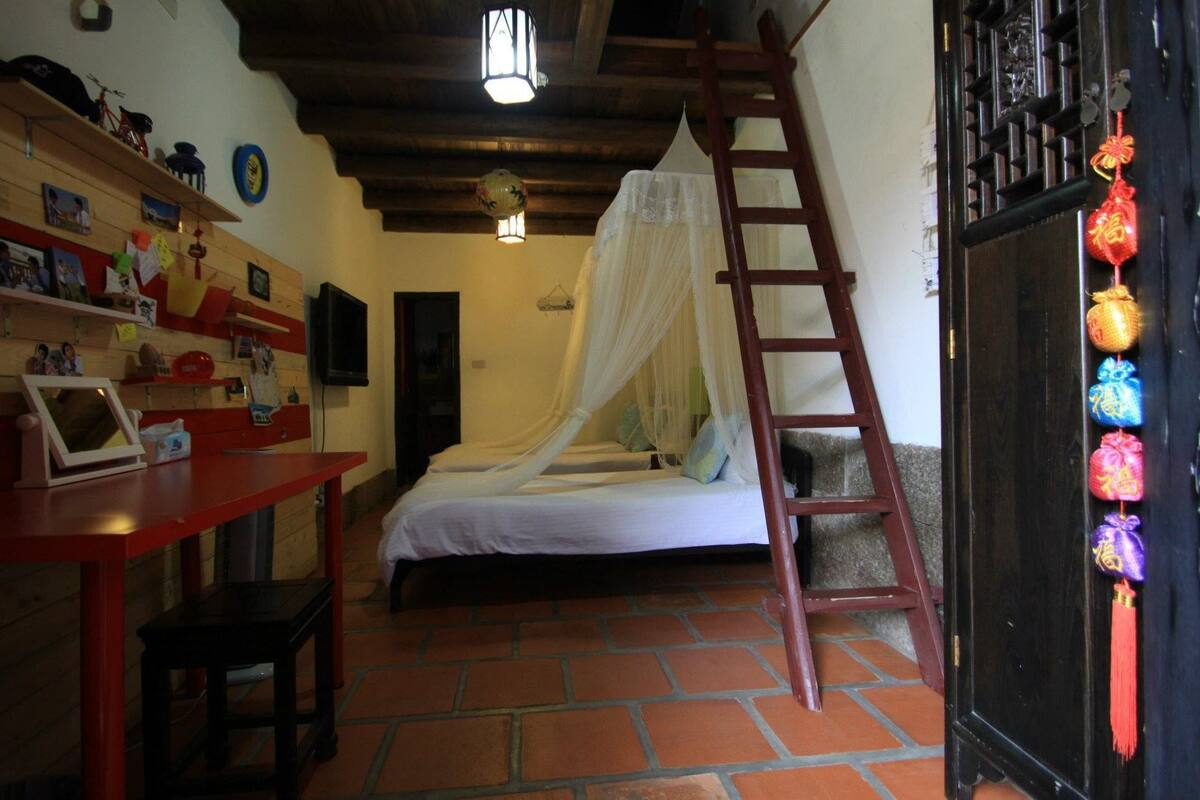 A bedroom with a bed under a white canopy, a wooden ladder, a red desk with a chair, and a shelf with various items.